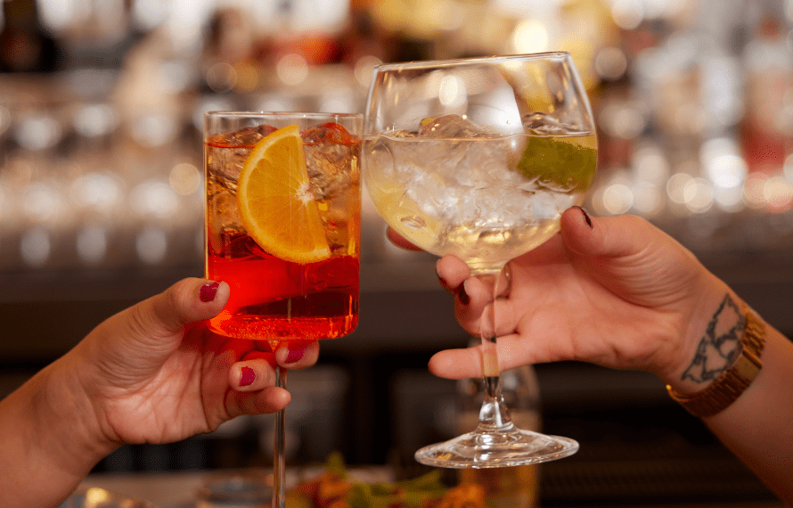 Two hands toasting with drinks in a bar setting, one holding a red cocktail with an orange slice, the other holding a clear drink with lime.
