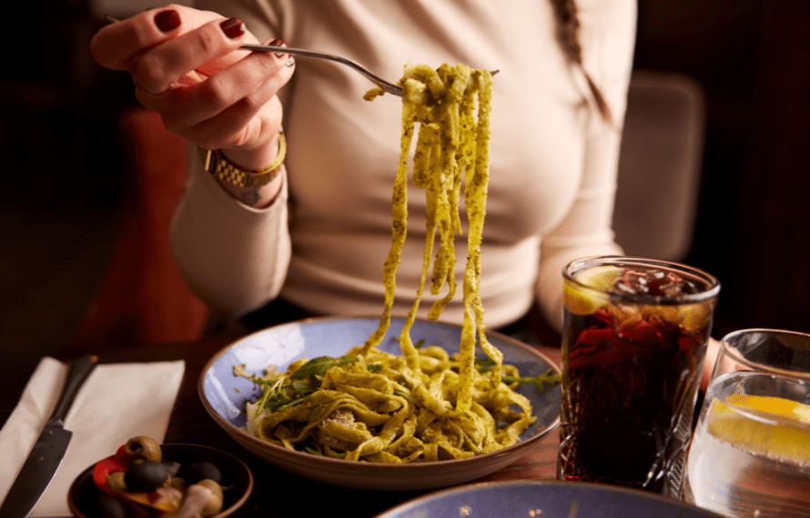 A person holding a forkful of green pesto pasta above a bowl of the same dish.