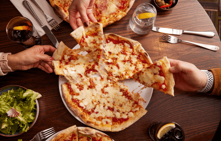 Three hands holding slices of cheese and tomato pizza over a table set with drinks