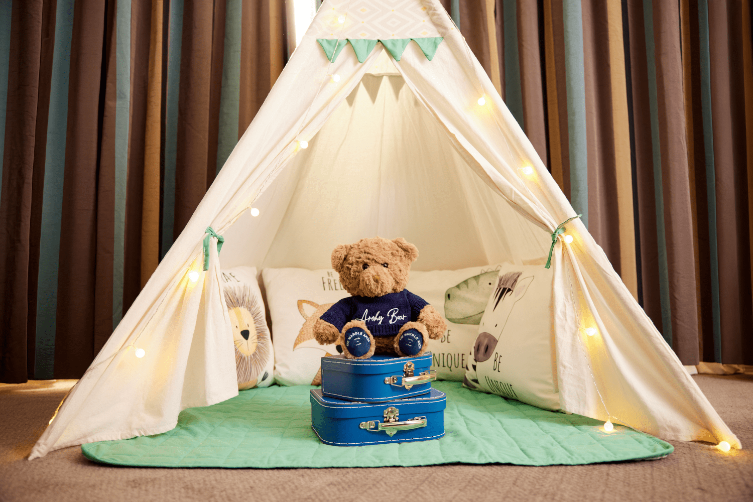 Children’s teepee play tent set up indoors with soft lighting and fairy lights, featuring animal‑print cushions and a teddy bear sitting on stacked blue suitcases on a padded green mat.