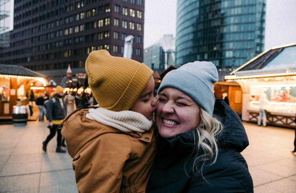 Child in a mustard hat kissing a smiling woman’s cheek, with illuminated market stalls behind them.